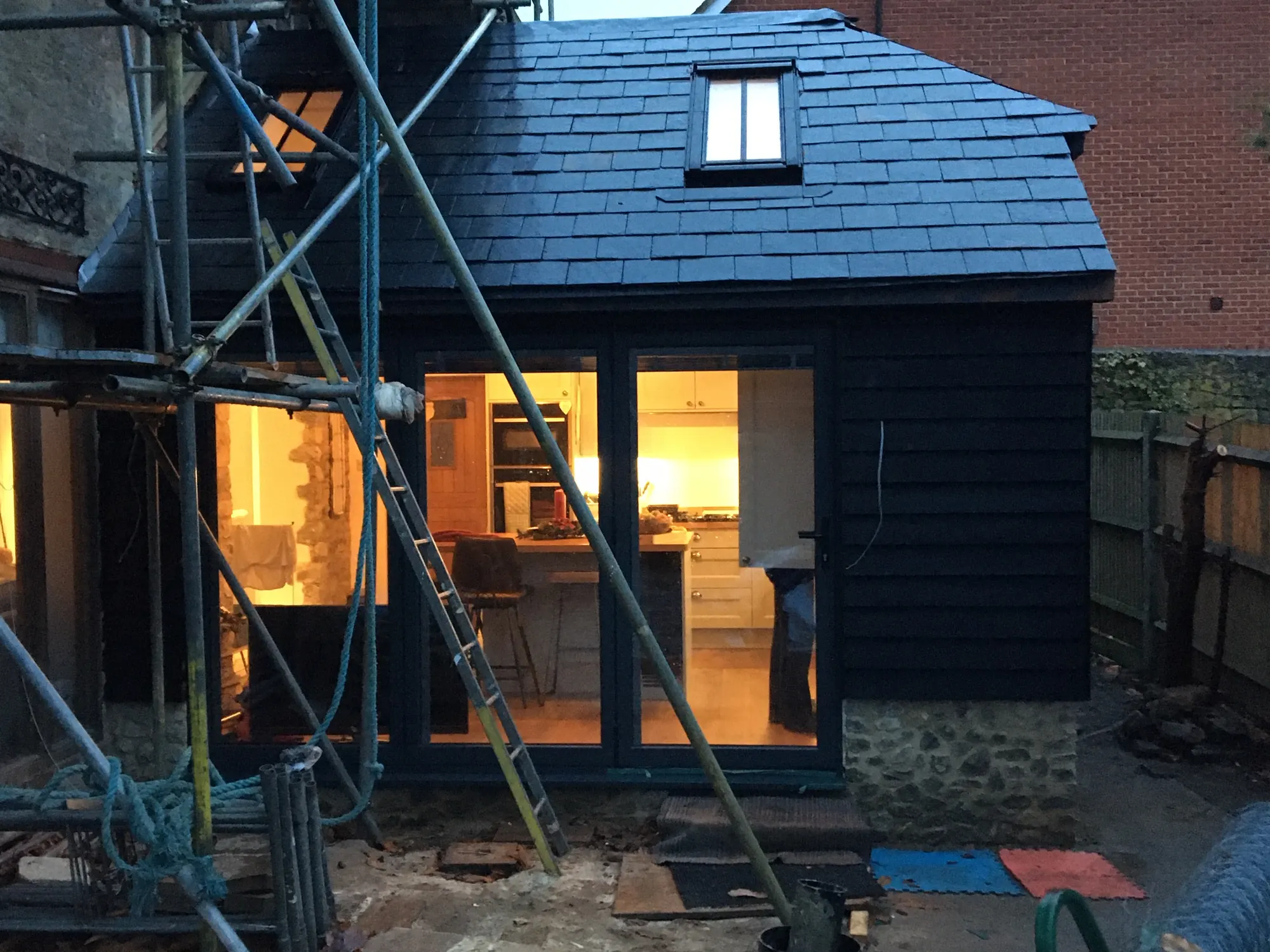 A newly slated extension at dusk with the scaffolding still standing and warm kitchen light coming through the glass doors