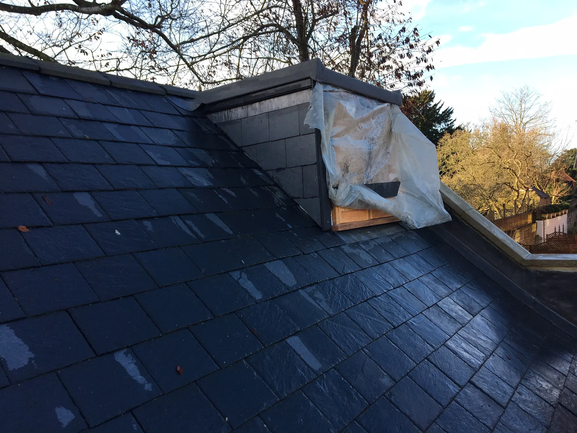 A fresh slate roof with the dormer wrapped in polythene and autumn trees behind