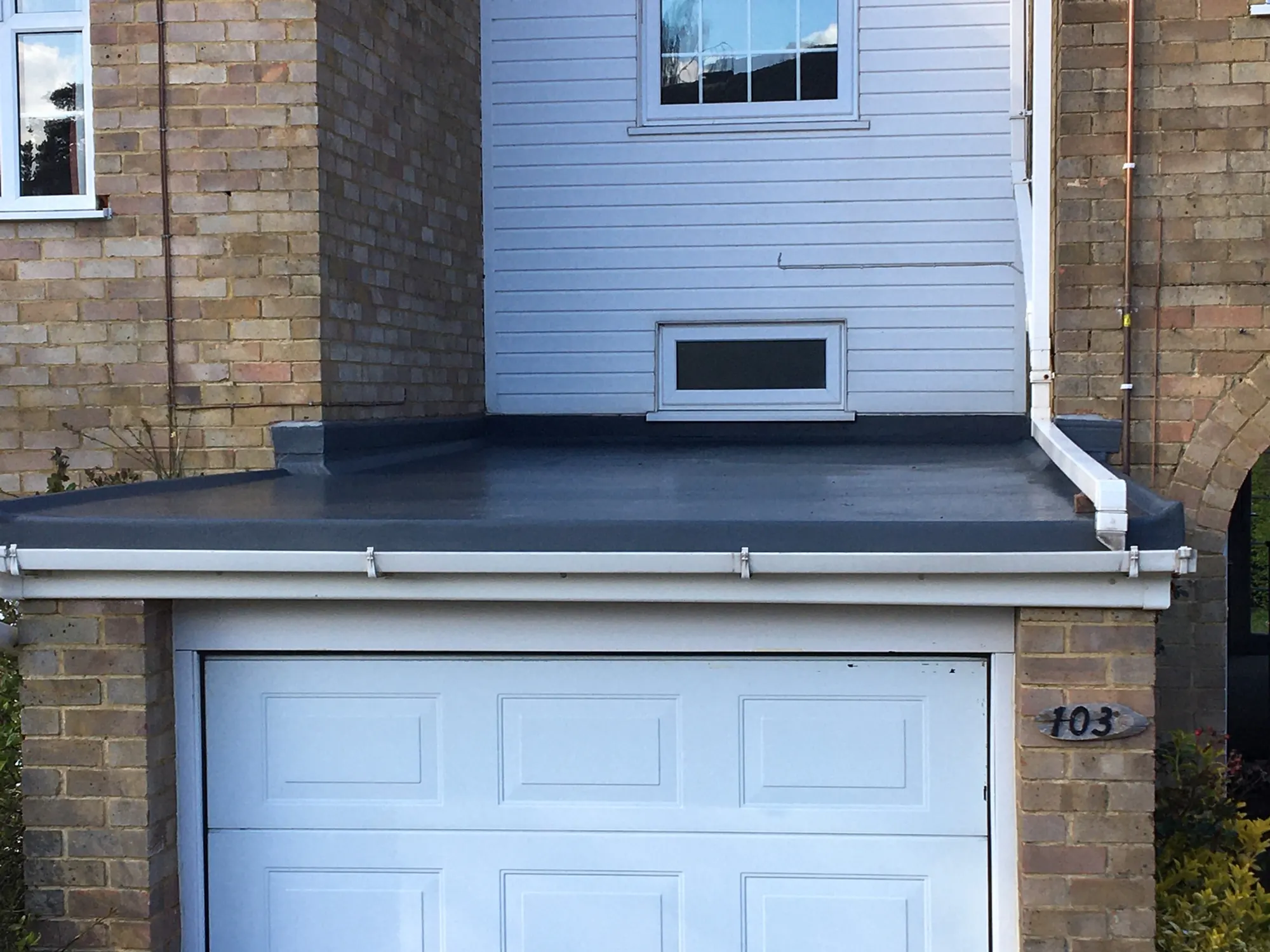 A grey flat garage roof with new fascia and downpipe above a white garage door